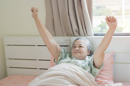Mujer asiática mayor acostada en su cama por la mañana bostezando con los brazos levantados en un estiramiento. Saludo nuevo día, buen concepto de la mañana