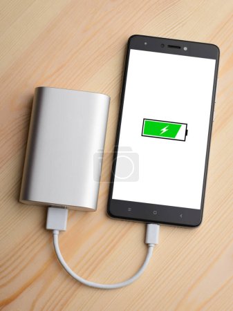 Modern smartphone lays on light wooden table while charging with power bank with quick charge. Illustration of green battery with lightning is on the mobile phone screen while charging
