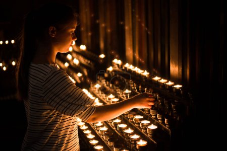 Religious woman putting burnng candle between many other church candles, praying for someone