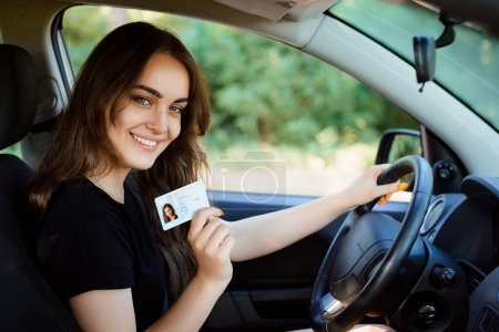 Foto de Sonriendo joven hembra con apariencia agradable muestra orgullosamente su licencia de conducir, se sienta en un coche nuevo, siendo joven conductor sin experiencia, se ve con expresión alegre - Imagen libre de derechos