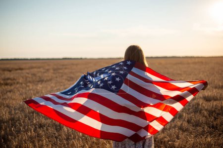 Téléchargez les photos : Jeune fille avec drapeau des États-Unis dans le domaine du seigle. Champ de céréales sans fin le soir avec une fille faisant une promenade avec un drapeau - en image libre de droit