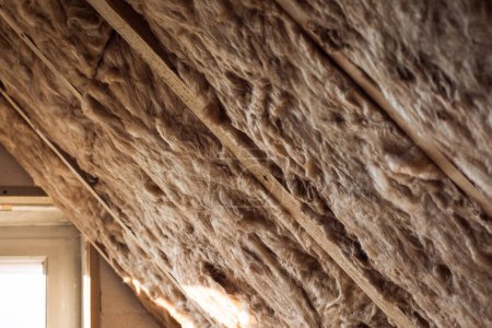 Téléchargez les photos : Laine de verre et mousse de polystyrène dans un cadre en bois sur un mur incliné près du plafond en bois et une fenêtre dans une maison privée. Chauffer les murs avec de la fibre de verre et de la mousse plastique. - en image libre de droit