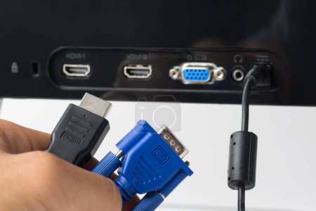 Foto de La mano del hombre sostiene cables HDMI y VGA contra un monitor con puertos. Elija entre una conexión HDMI moderna y una conexión VGA antigua - Imagen libre de derechos