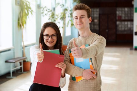Male and female students show thumbs up holding exercise books, notebooks and other learning materials in hall of their university looking happy to study in leading conventional university