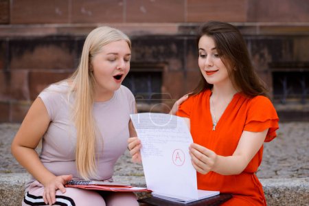 Photo for Two excited students looking at examination paper together sitting near university building in a desk. Concept of receiving the highest mark for examination or test - Royalty Free Image