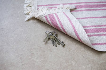 Grauer Schlüssel unter dem gestreiften weiß-rosafarbenen Teppich auf Laminatboden versteckt.