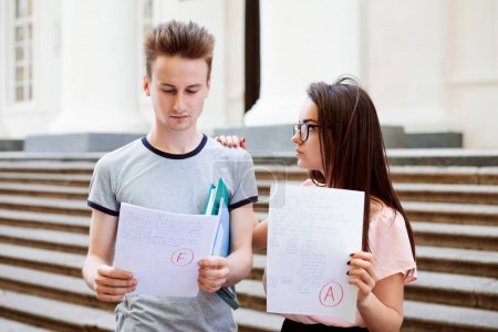Two students with test results. Female student have received excellent A grade for the test, but her male friend have failed. Girl looks sympathetically at her friend and holds hand on his shoulder