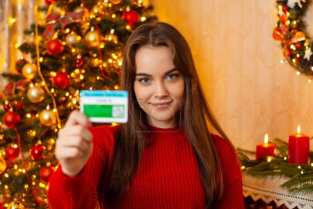 Photo for Pretty young girl with loose hair with Christmas tree on the background shows validated coronavirus certificate. Winter holidays, vaccine, covid-19 concept - Royalty Free Image