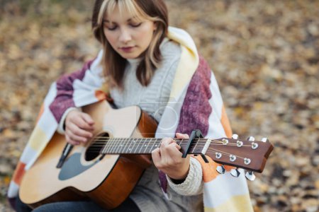 Photo for Young girl play the guitar outdoors. Smiling beautiful girl covered in warm plaid playing the guitar - Royalty Free Image