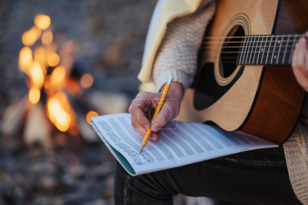 Photo for Girl composer playing the guitar composing new song outdoors near bonfire. Playing the guitar in autumn park in the evening, making new composition - Royalty Free Image