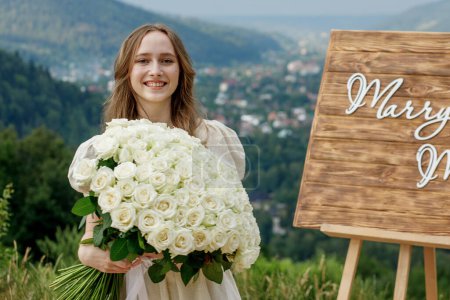 Photo for Girl holding a large bouquet of roses on the day of the proposal on the background of a decorative easel with the inscription MERRY ME and a mountain landscape. - Royalty Free Image