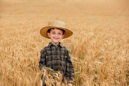 A smiling little farmer boy in a plaid shirt and straw hat poses for a photo in a wheat field. Heir of farmers.