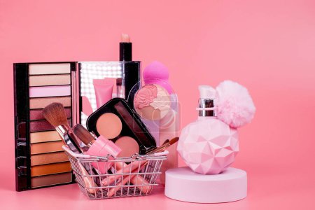 Foto de Concepto creativo con carrito de compras con maquillaje sobre un fondo rosa. Perfume, esponja, cepillo, rímel, sombra de ojos, brillo de labios en la cesta. - Imagen libre de derechos