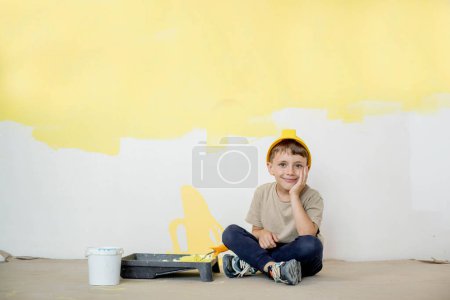Photo for A boy in a construction uniform sits near paint paints. Repairs. Painting the walls. - Royalty Free Image