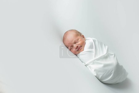 Téléchargez les photos : Un bébé de 3 jours enveloppé dans un tissu blanc dormant sur un fond blanc. - en image libre de droit