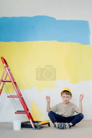 Photo for A boy in a construction uniform sits near paint paints. Repairs. Painting the walls. - Royalty Free Image