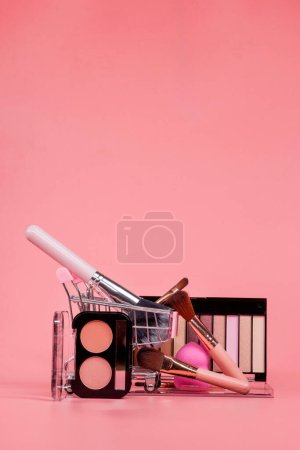 Téléchargez les photos : Concept créatif avec chariot d'achat avec maquillage sur fond rose. Parfum, éponge, pinceau, mascara, ombre à paupières, brillant à lèvres dans le panier. - en image libre de droit