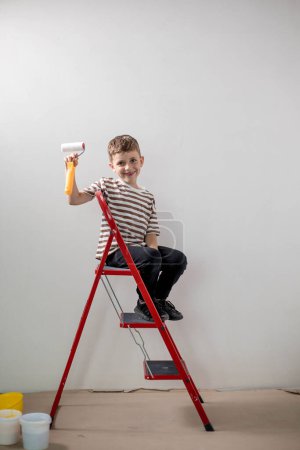 Photo for A child builder stands on a ladder and paints white walls in an apartment. A child with a paint roller during renovation. - Royalty Free Image