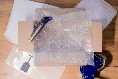 Open box with wrapped items, adhesive tape, scissors, paper and bubble wrap on wooden table, flat lay.