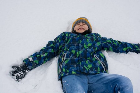 Photo for Cheerful boy lies in the snow. Active outdoors leisure for kids on nature in snowy winter day. - Royalty Free Image