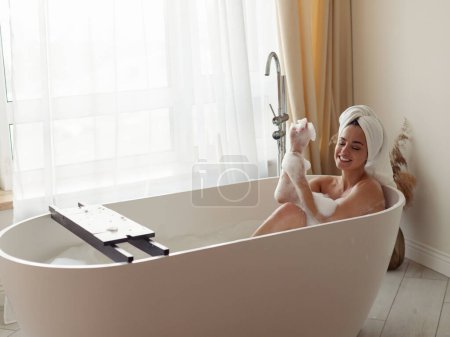 Joyful young Caucasian attractive woman lying in bathtub with foam naked with towel on head relaxing in bathroom enjoying process. Pretty smiling female enjoys morning hygiene body care routine
