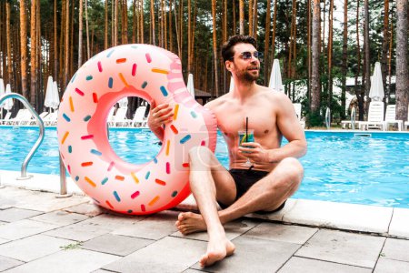 Téléchargez les photos : Homme souriant dans les lunettes de soleil profitant du cocktail tout en tenant anneau gonflable près de la piscine. Été, vacances, mode de vie concept - en image libre de droit