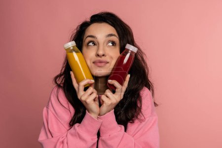 Portrait of a pensive brunette touching her cheeks with a pair of glass bottles filled with orange and tomato juice during the photo shoot