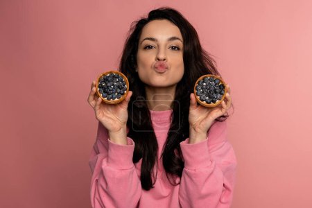 Photo for Young woman posing with a couple of appetizing fruit tartlets on the pink background during the studio photo shoot. Love of sweet food concept - Royalty Free Image