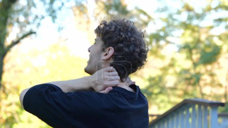 Photo for Back view young Caucasian man massaging his neck, shoulders , and back in green park between trees. Rearview sportsman with curly brown hair in black t-shirt feeling pain in his neck, shoulders. - Royalty Free Image