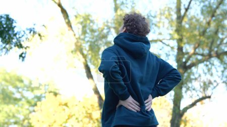 Photo for Rearview sportsman stretching body before working out. Back view Young Caucasian man massaging his back in green park between trees. - Royalty Free Image