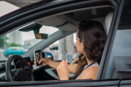 Photo for Smiling pretty lady making photo with driving license while using mobile phone in the car. Travel adventure drive, happy summer vacation concept - Royalty Free Image