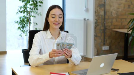 Photo for Caucasian young woman with dark long hair sitting on table in front of laptop and counting money. Happy business girl sitting at the table and counting money with pleasure, she is feeling success. - Royalty Free Image