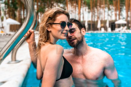 Photo for Smiling cute blonde woman in the bikini standing together with a young man in an outdoor swimming pool. Romance and summer vacation concept - Royalty Free Image