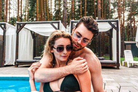 Téléchargez les photos : Beau jeune homme amoureux passionné embrassant son compagnon féminin attrayant près d'une piscine extérieure. Romance et vacances d'été concept - en image libre de droit