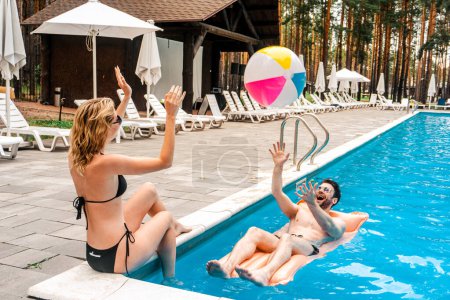 Schlanke Frau im Bikini wirft einen aufblasbaren Ball zu einem fröhlichen Mann mit Sonnenbrille, der auf der Luftmatratze im Freibad liegt