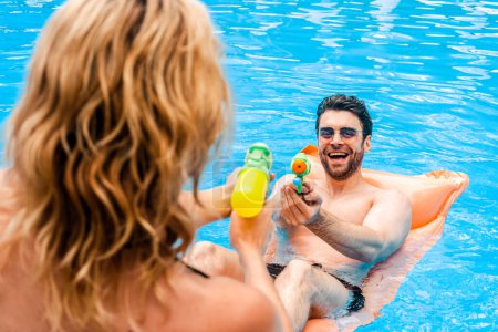 Photo for Mischievous guy in sunglasses lying on the air mattress in the outdoor swimming pool shooting his squirt gun at the blonde - Royalty Free Image