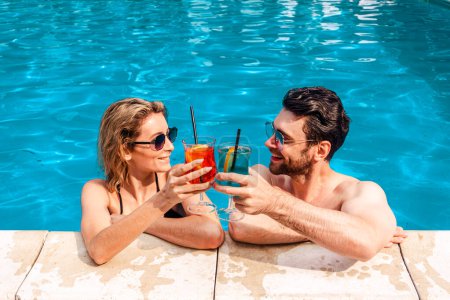 Photo for Smiling young woman and a cute dark-haired guy clinking glasses of cocktails in an outdoor swimming pool. Leisure and summer vacation concept - Royalty Free Image