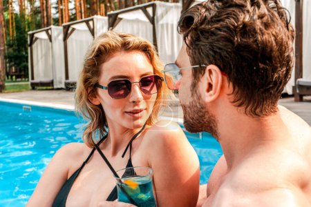 Photo for Portrait of a cute young lady with a glass of cocktail sitting next to her boyfriend at the holiday resort poolside. Romance and summer vacation concept - Royalty Free Image