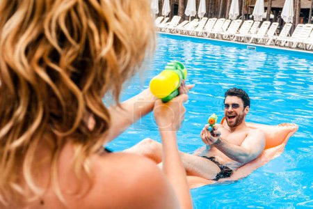 Blonde woman playing squirt guns with a cheerful young man in sunglasses lying on the air mattress in the outdoor swimming pool