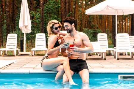 Photo for Smiling woman and her pleased boyfriend clinking glasses with cocktails while sitting on the swimming pool edge. Rest and relaxation concept - Royalty Free Image