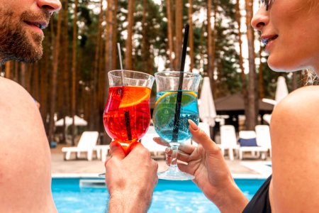 Photo for Cropped photo of a bearded guy and a woman in sunglasses clinking cocktail glasses while sitting at the poolside in the recreation area - Royalty Free Image