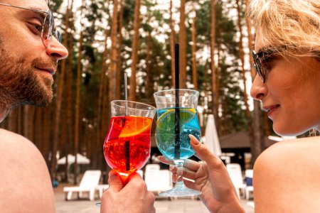 Retrato de una joven pareja romántica de turistas en gafas de sol sentados al aire libre tintinear vasos de cóctel mientras se miran entre sí