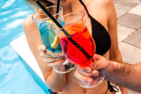 Cropped photo of a slim female in the bikini clinking cocktail glasses with a man by an outdoor swimming pool with the blue water