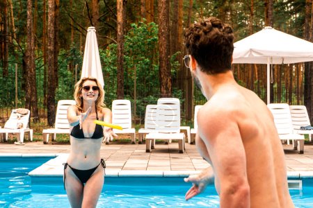 Photo for Cheerful blonde lady throwing a yellow disc to a dark-haired man standing in front of her in the outdoor pool. Summer vacation and active leisure concept - Royalty Free Image