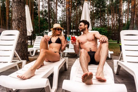 Photo for Happy couple of holidaymakers in sunglasses lying on the white loungers and clinking glasses with colorful cocktails. Leisure and summer vacation concept - Royalty Free Image