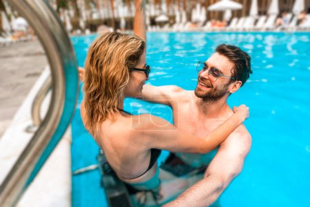 Photo for Smiling slim blonde female in the bikini hugging her cheerful young man in the outdoor swimming pool. Romance and summer vacation concept - Royalty Free Image