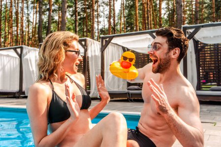Photo for Merry guy holding a small inflatable duck toy in front of his pleased girlfriend seated with him at the poolside - Royalty Free Image