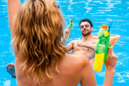 Blonde Frau steht vor einem zufriedenen jungen Mann mit einer Spritzpistole in der Hand, die auf der Luftmatratze im Schwimmbad liegt
