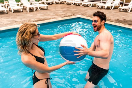 Photo for Cheerful dark-haired man and a blonde woman in the bikini pulling the ball in different directions while standing in the outdoor swimming pool - Royalty Free Image