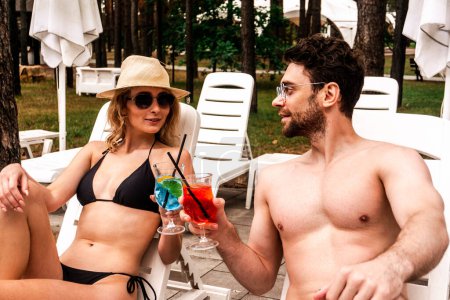 Photo for Slim woman in the bikini and a young man in sunglasses clinking glasses with refreshing drinks while resting on the white plastic loungers outdoors - Royalty Free Image